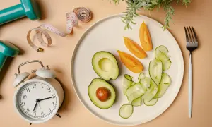 Zegar odmierza czas na zdrowy posiłek. Awokado, pomidory i ogórek na talerzu, obok miarki i hantli – to wszystko symbolizuje dietę imitującą post.