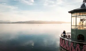 Ein Paar genießt den Sonnenuntergang am Leuchtturm Lindau. Der Bodensee glänzt, Berge im Hintergrund.