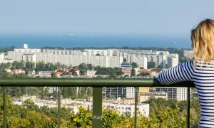 Widok z punktu widokowego w Oliwie na morze i osiedle. Kobieta w bluzce w paski podziwia panoramę.