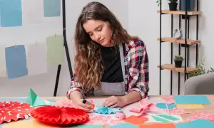 Chica creando manualidades para niños de 12 a 15 años. Pliegues de papel, abanicos y adornos coloridos sobre la mesa.