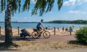 Rowerzysta z przyczepką jedzie ścieżką przy plaży nad Pogorią. Ludzie odpoczywają na piasku.