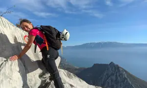 Donna sorridente su roccia con zaino e casco, vista lago e montagne. Un facile sentiero sul monte Pizzocolo.