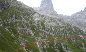 Sendero marcado en rojo asciende por la ladera verde hacia el imponente Sotres Urriellu, una cumbre rocosa en los Picos de Europa.