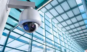 Caméra de surveillance fixée à un mur, prête à filmer. Le bâtiment moderne avec sa façade vitrée reflète le ciel bleu.