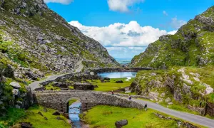 Eine malerische **irland landschaft** mit einem steinernen Bogenbrücken über einem Bach, der in einen See mündet, umgeben von grünen Hügeln und felsigen Bergen.