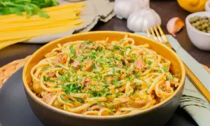 Eine Schüssel mit Thunfisch Pasta, garniert mit Petersilie und Zitronenschale. Daneben liegen Spaghetti, Knoblauch und Kapern.