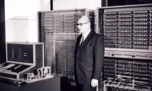 Mężczyzna w garniturze stoi obok ogromnej maszyny z mnóstwem przełączników i lamp. To prawdopodobnie pierwszy komputer na świecie, przełomowa technologia.