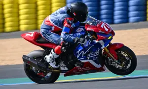 Motocyklista w niebiesko-czerwonym kombinezonie na torze wyścigowym, z tyłu żółte i niebieskie bandy.