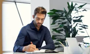 Hombre concentrado escribiendo en un diario en su escritorio, rodeado de plantas y un portátil.