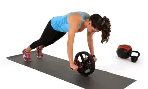 Kvinna utför ab wheel rollout på en träningsmatta. En kettlebell och en medicinboll syns i bakgrunden.
