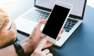 Dłoń trzyma biały smartfon, który nie chce się włączyć. Obok leży laptop, a w drugiej dłoni kubek z kawą.