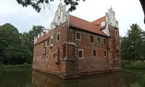 Zamek na wodzie w Wojnowicach, otoczony zielenią, z ceglanymi murami i charakterystycznymi białymi szczytami.
