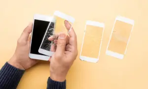 Ręce trzymają biały telefon z pękniętym szkłem hartowanym 5d. Obok leżą dwa inne telefony z rozbitymi ekranami.