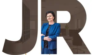 Kobieta w niebieskiej marynarce na tle liter JR i wnętrza biura.
