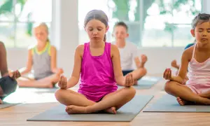 Niños practicando ejercicios de respiración y meditación en un salón de clases, sentados en colchonetas con los ojos cerrados.