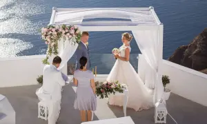 Piękny ślub w Grecji. Para młoda stoi pod białym baldachimem, w tle błękitne morze. Ceremonia odbywa się na tarasie z widokiem na ocean.