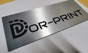 Metalowa tabliczka grawer z logo "Dor-Print". Wygląda jakby była wykonana z aluminium.