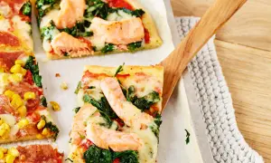 Pizza mit Lachs und Spinat, belegt mit geschmolzenem Käse, wird mit einem Holzschieber serviert.