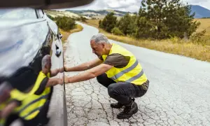 Hombre con chalecos reflectantes, agachado junto a un coche averiado en una carretera rural.