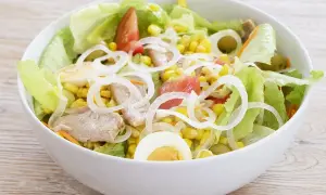 Deliciosa ensalada mixta con atún, huevo duro, maíz, tomate y cebolla, servida en un cuenco blanco sobre una mesa de madera.