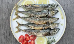 Cinq sardines au four, dorées et savoureuses, servies avec des quartiers de citron, des tomates cerises et des herbes fraîches sur une assiette blanche.