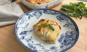 Porzione di lasagne di zucchine gratinate su piatto decorato, con basilico fresco e una teglia sullo sfondo.