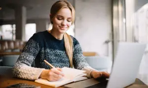 Uśmiechnięta studentka notuje w zeszycie, korzystając z laptopa. Cieszy się z możliwości, jakie dają jej zniżki studenckie.