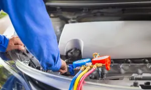 Mechanik w niebieskim kombinezonie wykonuje samodzielne nabijanie klimatyzacji w samochodzie, używając manometrów i kolorowych węży.