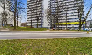 Dwa wysokie bloki mieszkalne z balkonami, ulica z pasami i zielony trawnik.