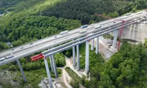 Widok z lotu ptaka na budowę betonowego mostu nad lasem, z ruchem samochodowym na ukończonej części.