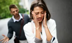 Una donna piange disperata, con le mani sul viso, mentre un uomo alle sue spalle gesticola con frustrazione. L'immagine cattura un momento di profonda infelicità di coppia.