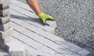 Dłoń w rękawiczce układa kostkę brukową na podsypce.