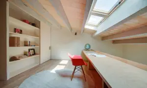 Un pequeño patio de luz interior baña con sol un escritorio de madera, una silla roja y una estantería.