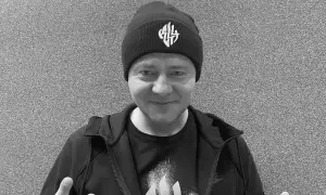 Mężczyzna w czapce z logo, w bluzie i koszulce. W tle szara ściana. Nie wiadomo, **joka na co zmarł**.