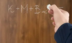 Dłoń pisze kredą na tablicy: K+M+B. Nagle przychodzi epifania, rozwiązanie problemu.