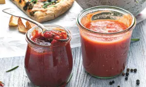Dwa słoiki domowego sosu do pizzy, jeden z łyżką pełną aromatycznej masy. Obok kawałek pizzy i przyprawy.