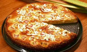 En saftig rabarberkladdkaka med smulad vit choklad och färska rabarberstjälkar i bakgrunden.