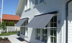 Grå markiser monterade på ett vitt trähus ger skugga åt fönstren. En vacker trädgård med stenläggning och staket syns i bakgrunden.