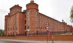Imponujący, ceglany **księży młyn** w Łodzi, z charakterystycznymi wieżyczkami i długim rzędem okien. Obok znaki drogowe.