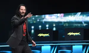 Keanu Reeves na scenie, wskazując palcem, w koszulce z logo Cyberpunk 2077. Gra zarobiła miliony, a on jest jej twarzą.