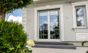 Eleganckie okno bez klamki z białymi szprosami, odbijające błękitne niebo i chmury. Obok zielone krzewy i biała róża.