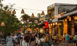 Bunte Lichter und Menschenmassen beleuchten die **Hoi An Altstadt**. Ein vietnamesisches Banner weht über einer belebten Straße voller Geschäfte und Restaurants.