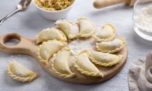 Gotowe do gotowania pierogi ruskie ułożone na desce, posypane mąką. Obok miska z farszem i wałek.