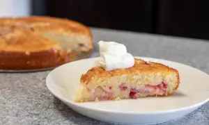 En bit saftig sockerkaka med rabarber, toppad med en klick grädde. Hela kakan syns i bakgrunden.