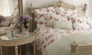 En säng med blommig shabby chic inredning, prydd med rosor och lavendel. Ett runt spegelbord med en vas full av blommor.