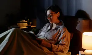 Kobieta w okularach czyta książkę w łóżku przy lampce nocnej. Czy czytanie po ciemku psuje wzrok?