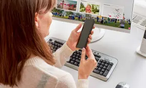 Kobieta trzyma telefon, szukając informacji, jak podłączyć telefon do monitora. Na ekranie komputera widać zdjęcia.