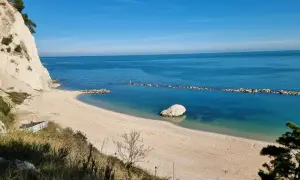 Spiaggia dorata e mare cristallino sulla Riviera del Conero, con una scogliera bianca a picco sull'acqua e un grande masso solitario.