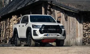 Biały Toyota Hilux GR Sport z czarnymi dodatkami, zaparkowany przed drewnianym budynkiem z polanami drewna.