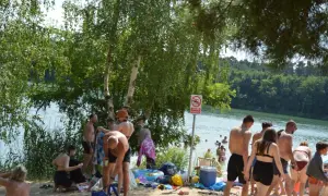 Ludzie odpoczywają nad jeziorem, niektórzy kąpią się, inni siedzą na plaży. W tle widać las. To idealne miejsce na relaks, jakby czas się zatrzymał, niczym w starożytnym rydwanie.